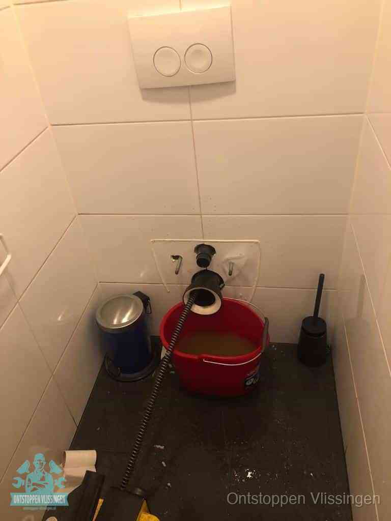 Een ontstopte wand-wc met verwijderde toiletpot waar een ontstoppingsveer in de wanduitgang verdwijnt, het achterhalen waar de verstopping zit in Vlissingen