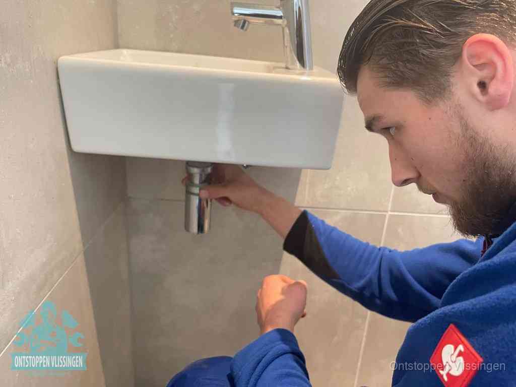 Een monteur draait met de hand de zwanenhalssifon onder een fonteinwasbak in de toiletruimte los om de bron van rioollucht in Vlissingen te vinden