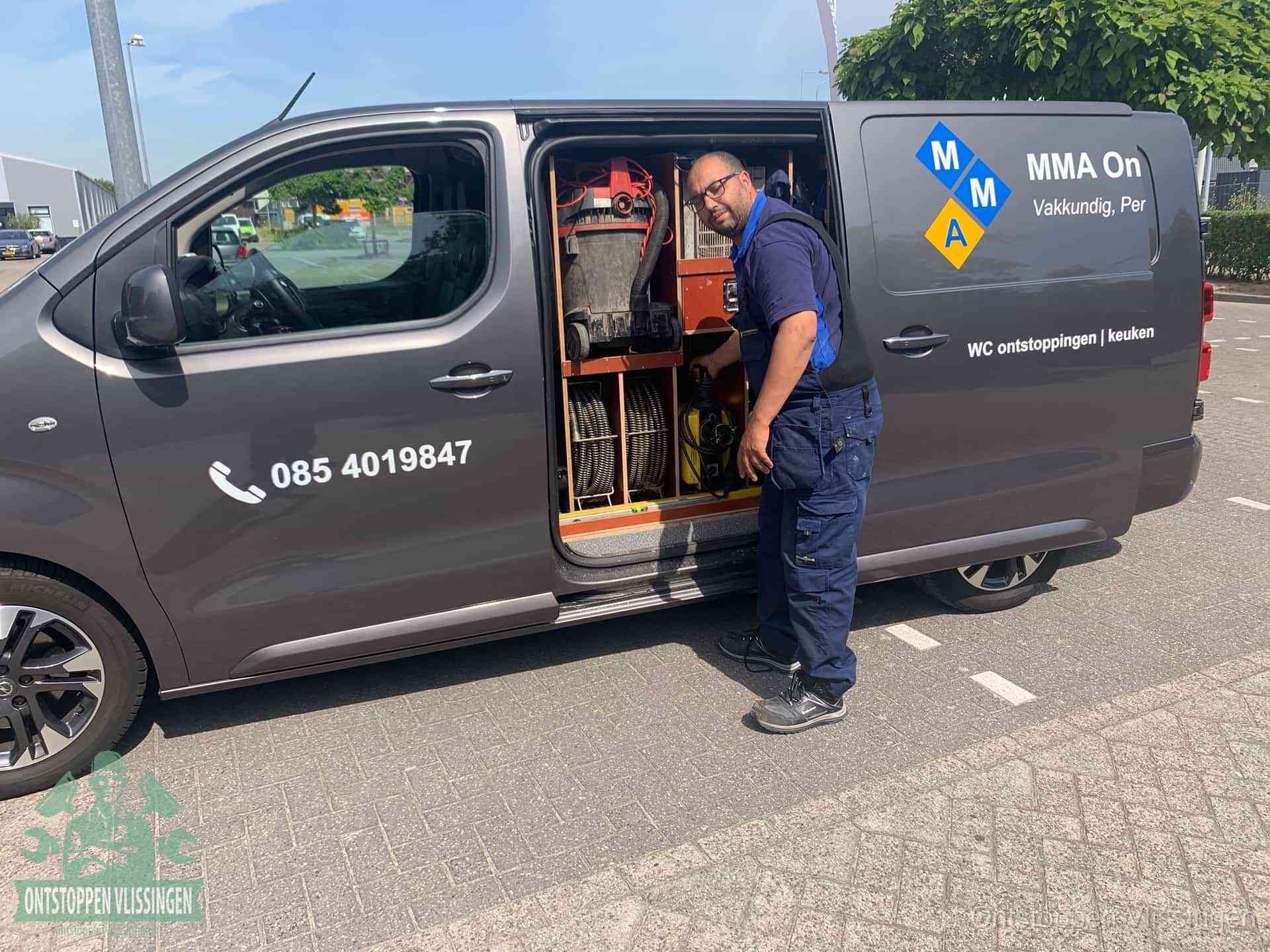 Een monteur bij de open schuifdeur van zijn bedrijfsbus op een parkeerplaats, klaar om de oorzaak van terugkerende afvoerverstoppingen aan te pakken in Vlissingen