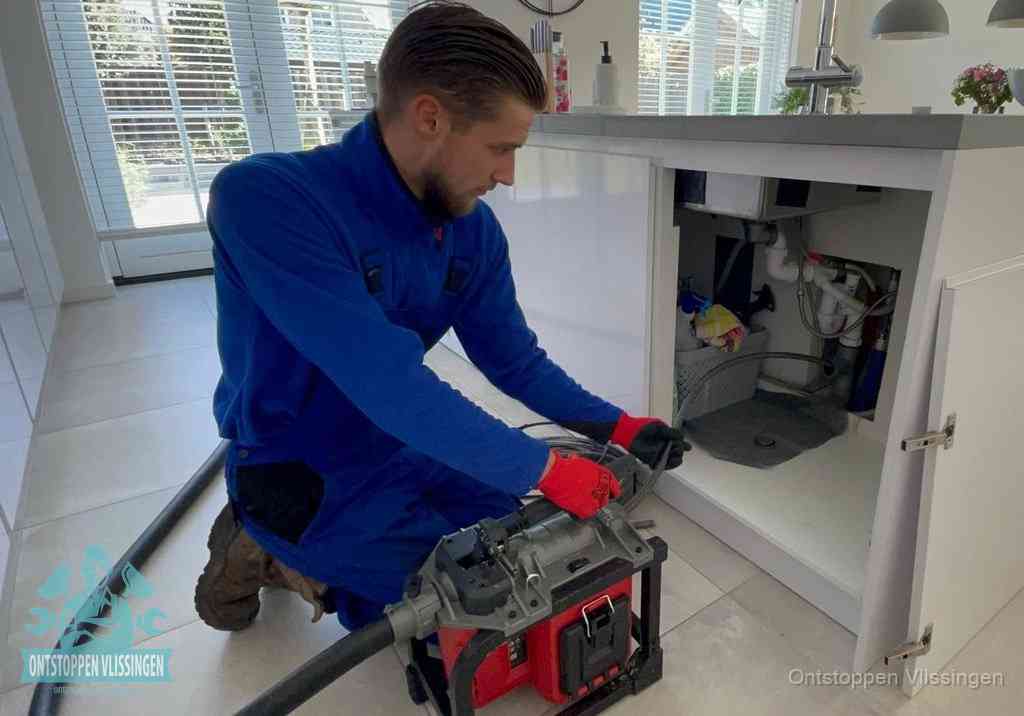 Een monteur voert een professionele elektrische ontstoppingsmachine in de afvoer onder de spoelbak tijdens het gootsteen ontstoppen in Vlissingen